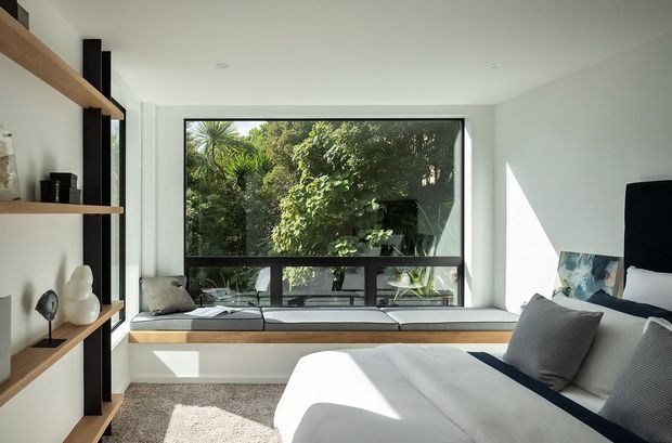 Bright contemporary bedroom with a built-in window seat overlooking lush greenery. The room features a large black-framed window, white walls and bedding, a warm timber open shelving unit, a deep charcoal structural or shelving accent, and a soft neutral carpet. Natural light creates a calm, airy atmosphere.