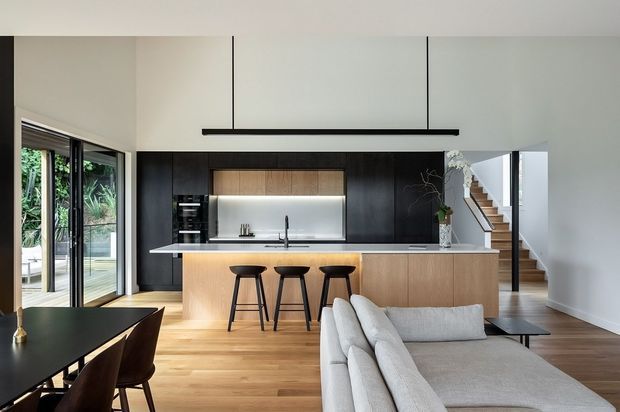Open-plan contemporary kitchen and living area with a timber island, dark cabinetry, integrated appliances, pale stone benchtops, and three black bar stools. A large light-grey sectional sofa sits in the foreground beside a dining table, while full-height glazing opens toward an outdoor area and a stairway is visible at the rear.