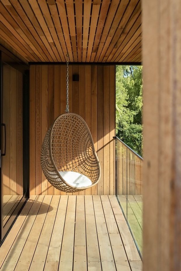 A warm timber-lined outdoor or semi-enclosed balcony terrace with timber decking, slatted timber ceiling, vertical timber wall cladding, and a suspended woven hanging chair. A glass balustrade opens toward leafy vegetation, creating a private, sunlit retreat.