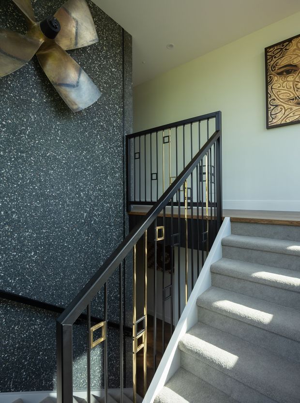 Interior view of a residential stairway with light grey carpeted steps, black metal balustrades, and a dark speckled feature wall. Gold-toned geometric detailing appears within the stair railing, while a large metallic ceiling fan and decorative wall artwork add visual interest. The space is bright and contemporary with a dramatic, high-contrast palette.