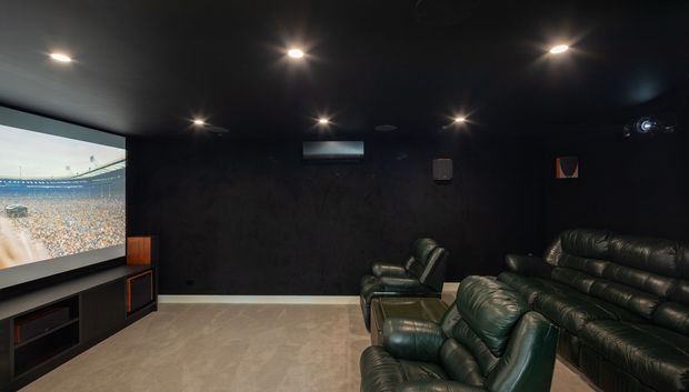 A dark, dedicated home theatre with black walls and ceiling, recessed downlights, a large projection screen displaying a sports crowd, and multiple rows of plush black leather recliner seating. The room has a light neutral carpeted floor and built-in audiovisual cabinetry beneath the screen.