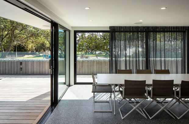 A bright contemporary dining area opening directly onto a timber deck through expansive floor-to-ceiling glazing and folding glass doors. A long rectangular dining table with brown upholstered chairs occupies the room, with sheer dark curtains along the glazed wall. An outdoor spa or pool structure and leafy garden are visible beyond the deck.