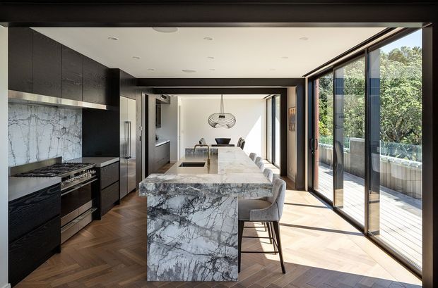 A refined contemporary kitchen and dining space viewed from the living area. The room features a large marble-look island with waterfall ends and integrated seating, dark full-height cabinetry, a matching stone splashback, and a stainless-steel cooking range. Warm herringbone timber flooring contrasts with the black joinery and pale stone. Full-height glazed sliding doors open to a timber deck and leafy outdoor setting, while a sculptural pendant hangs above the dining table in the background.