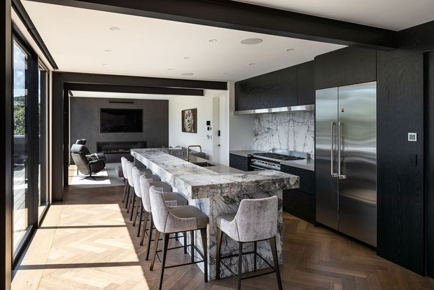 A refined open-plan kitchen with a long marble island and integrated seating for several people. Dark timber or black cabinetry spans the back wall and surrounds a stainless-steel refrigerator and cooking area, with a marble backsplash. Warm herringbone timber flooring contrasts with the dark joinery, while full-height glazing along the left brings in abundant daylight. A media or living area is visible beyond the kitchen.