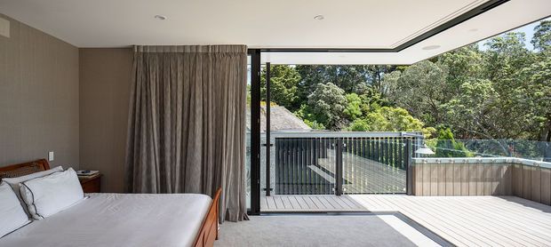 Contemporary bedroom opening directly onto a spacious timber deck or balcony through full-height glazed sliding doors. The room has a low-profile bed with white linens, warm taupe textured walls, full-height grey-beige curtains, recessed ceiling lights, and dark-framed glazing. The outdoor area includes a glass balustrade, dark vertical screening, and a raised timber-clad edge, with dense greenery beyond.