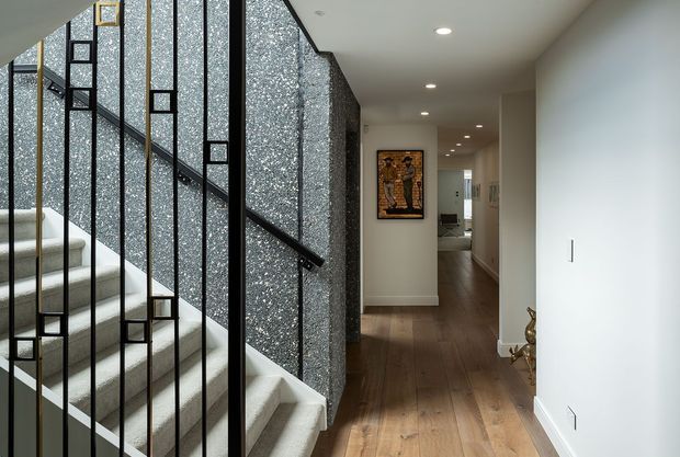 Interior view of a refined residential hallway and stairway. A curved carpeted staircase is enclosed by a striking black metal balustrade with geometric rectangular detailing, beside a textured dark grey feature wall. The hallway extends toward the rear of the home with warm timber flooring, recessed ceiling lights, white walls, and framed artwork.