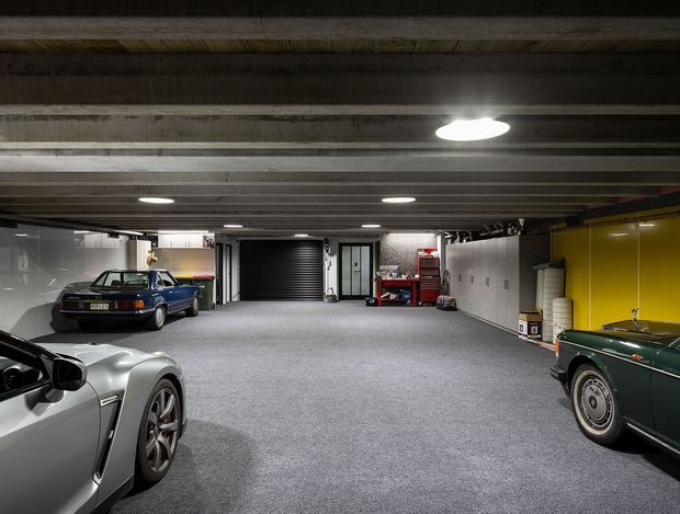 A large, low-ceilinged automotive garage or car showroom with several classic and contemporary cars parked on a grey carpeted floor. The space features exposed concrete beam ceiling construction, recessed circular lighting, white and bright yellow wall panels, a dark roller garage door, workshop storage, and a red tool cabinet. The image has an industrial, utilitarian character with high vehicle display capacity.