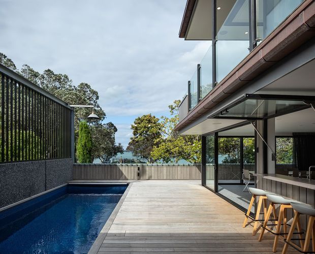 A contemporary multi-level home opening onto a timber pool deck and narrow swimming pool. The image shows extensive floor-to-ceiling glazing, dark slender structural frames, an outdoor bar or kitchen with stools beneath a covered overhang, upper-level glass balustrades, and a landscaped coastal-looking setting beyond the boundary wall.