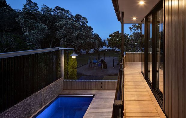 Twilight view of a modern elevated outdoor deck and narrow swimming pool beside a house. The deck uses warm timber boards and is bordered by dark vertical screening and metal-framed full-height glazing, with covered soffit lighting and a landscaped, tree-lined setting beyond.