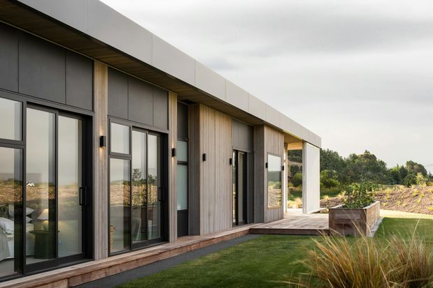 Contemporary single-storey residential facade viewed from a landscaped outdoor area, with strong horizontal roof overhangs, dark framed floor-to-ceiling glazing, vertical timber cladding, and a sheltered timber deck. The building opens toward a grassy garden and planted landscape under an overcast sky.