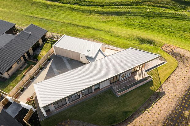 Aerial view of a contemporary single-storey residential property in a rural setting. The main house has an elongated low-pitched metal roof, extensive glazing, timber-toned exterior walls, and a covered deck or verandah. A separate flat-roofed outbuilding, paved courtyard, fencing, lawns, and adjoining cultivated fields are visible.