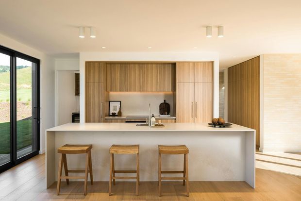 Bright contemporary kitchen featuring a large white island with seating for three, integrated timber cabinetry, open shelving, concealed appliances, and warm timber flooring. Black-framed glazing on the left brings in views of the surrounding landscape, while a light brick feature wall is visible at the right.