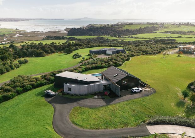 Aerial view of a contemporary rural property set among expansive green fields, landscaped grounds, and a curved asphalt driveway. The foreground house combines dark metal-clad forms with a steep gabled roof and a rounded or curved wing. Additional outbuildings are visible behind it, while a broad coastal inlet or shoreline and distant settlement appear in the background.