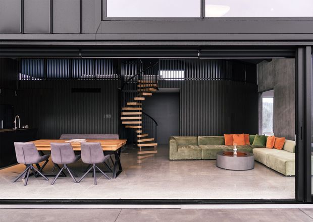 Open-plan contemporary living and dining area viewed through a large glazed opening. The space features dark vertically ribbed wall panels, a sculptural floating timber staircase, a long timber dining table with upholstered chairs, and a large L-shaped sectional sofa around a round coffee table. Concrete-toned flooring and extensive dark-framed glazing create an industrial, minimalist atmosphere.