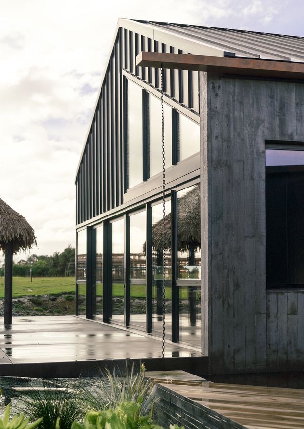 Exterior architectural photograph of a contemporary, angular building with a steeply sloped corrugated metal roof and vertically ribbed dark metal cladding. Full-height glazing forms the lower facade, while a raw concrete volume projects at the front. A timber-toned deck and covered outdoor structure with a rain chain sit beside landscaped grounds and a shallow water feature.