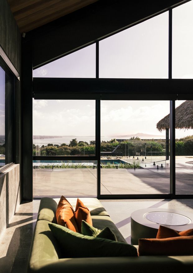 A dramatic, sunlit living room with a deep green sofa and orange cushions in the foreground, facing extensive black-framed floor-to-ceiling glazing. Through the windows is a terrace, swimming pool, landscaped garden, thatched outdoor structure, and a broad coastal or waterfront view. The room has a dark ceiling and wall framing, warm timber ceiling boards, and a polished concrete floor, creating a strong indoor–outdoor connection.