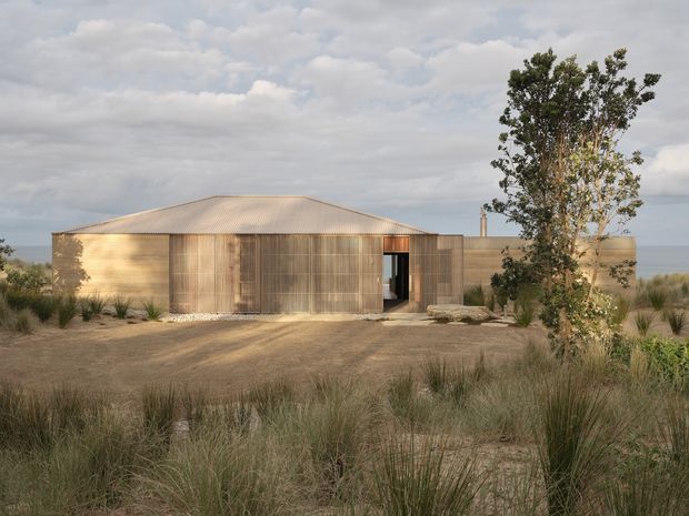 A low, single-storey rural building set within a natural grassland landscape. The structure has a simple elongated form, a shallow pitched corrugated metal roof, warm timber-clad walls, and a screened timber entry volume. Sparse native-style planting and sandy gravel ground surround the building, with a cloudy sky and distant open landscape beyond.