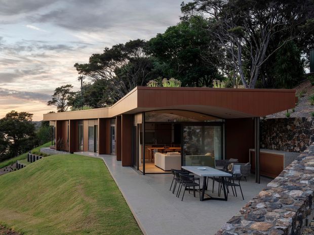 A low-slung contemporary house set into a sloping, heavily vegetated site. The building has a broad flat roof with warm rust-brown cladding, expansive glazed openings, and a curved covered outdoor terrace. An outdoor dining setting sits on a concrete patio beside a manicured lawn, with a rugged stone retaining wall rising along the right edge. Interior living and dining areas are visible through the glazing.