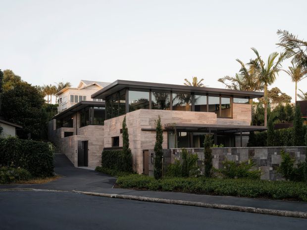 Street-facing view of a contemporary multi-level residence with strong horizontal lines, extensive dark-framed glazing, pale stone masonry walls, and flat roof forms. Dense hedging and tropical planting screen the property, with a driveway and low stone boundary wall visible in the foreground.