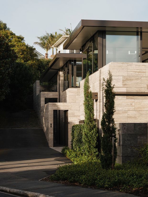A contemporary multi-level residential facade viewed from the driveway, with extensive pale stone wall cladding, dark-framed floor-to-ceiling glazing, projecting flat roof planes, recessed balconies, and a dark paved approach. Tall, narrow landscaped shrubs soften the strongly geometric building envelope.