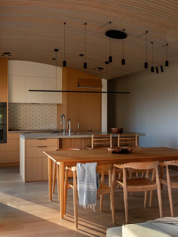 Warm contemporary open-plan kitchen and dining area with extensive light timber detailing. The kitchen features white flat-panel overhead cabinetry, timber lower cabinetry and pantry doors, a grey island benchtop, patterned geometric tiled splashback, and a timber-lined ceiling with multiple suspended black pendant lights. A large timber dining table and matching wooden chairs occupy the foreground, with soft afternoon light creating a calm, refined atmosphere.
