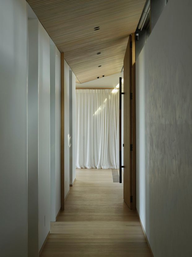 A narrow, softly lit residential hallway with pale timber flooring and a matching slatted timber ceiling. White walls and full-height vertical wall panels line the corridor, with warm timber door frames and a partially open door on the right. The hallway leads toward a softly draped, light-filled room, creating a calm and minimalist atmosphere.