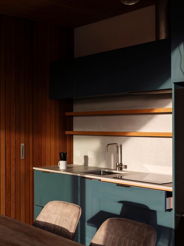 Compact, moody kitchen interior with deep teal lower cabinetry, a pale stone benchtop and splashback, open timber display shelves, and a dark overhead cabinet. The kitchen is framed by rich vertical timber wall panelling and a matching timber ceiling, with a stainless steel sink and gooseneck tap. Strong sunlight creates dramatic shadows across the cabinetry and work surface; the foreground includes part of a dark dining table and upholstered dining chairs.