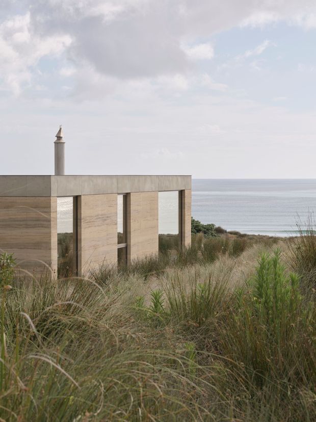 A minimalist coastal building set among tall dune grasses, with a low rectangular concrete or stone-clad structure, deep vertical openings, and a slender chimney. The calm ocean is visible directly behind the building under an overcast sky. The composition emphasizes the relationship between the restrained architecture and the natural coastal landscape.