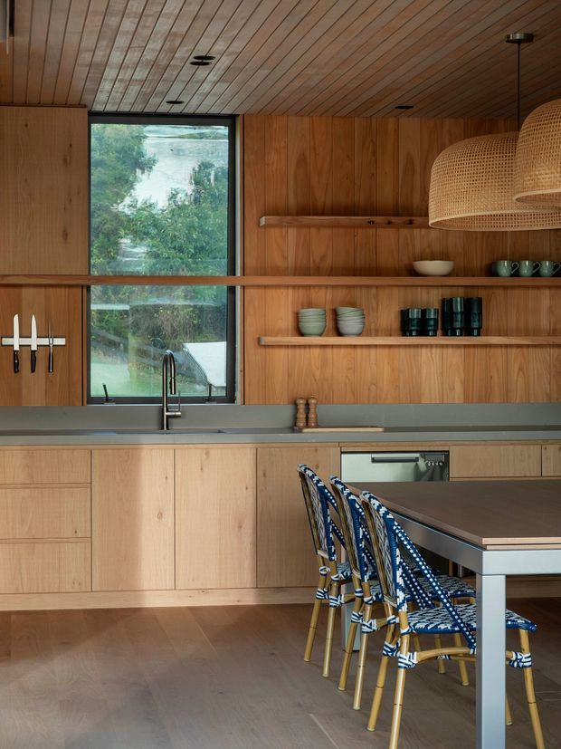 Warm, wood-lined contemporary kitchen with full-height timber cabinetry and open shelving, a long grey benchtop with integrated sink, black-framed window, stainless-steel tap and integrated oven. A dining table with patterned woven chairs occupies the foreground, while a large woven pendant light hangs over the dining area. The room has a strong timber ceiling and timber flooring, with a leafy outdoor view through the window.