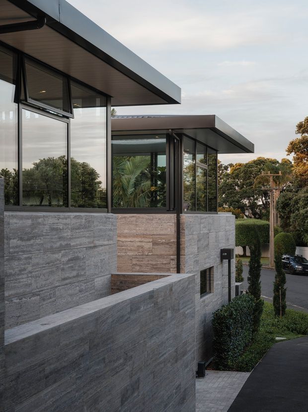 Exterior view of a contemporary multi-level residence on a sloping suburban site. The building features strong horizontal flat roof planes, extensive dark-framed floor-to-ceiling glazing, and textured grey stone or concrete wall surfaces. Layered terraces and retaining walls step down toward a landscaped driveway and street, with mature trees visible beyond.