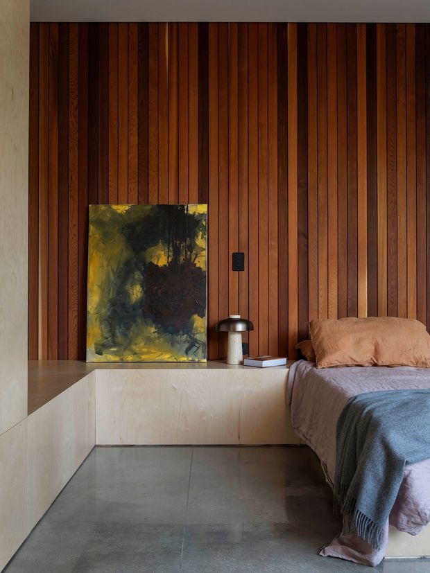 A warm, moody bedroom with a full-height vertical timber slat feature wall, pale built-in platform cabinetry and bedside ledge, polished grey concrete flooring, and a partially visible bed dressed in muted taupe and grey textiles. An abstract artwork and compact table lamp sit on the ledge, creating a restrained, contemporary interior.