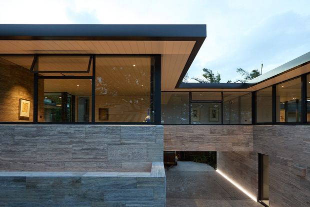 Modern low-profile residential exterior with extensive floor-to-ceiling glazing, dark metal framing, warm timber soffits, and horizontally laid grey stone cladding. The view looks across a sunken courtyard or driveway passage, with a covered connection between glazed wings and integrated linear lighting.