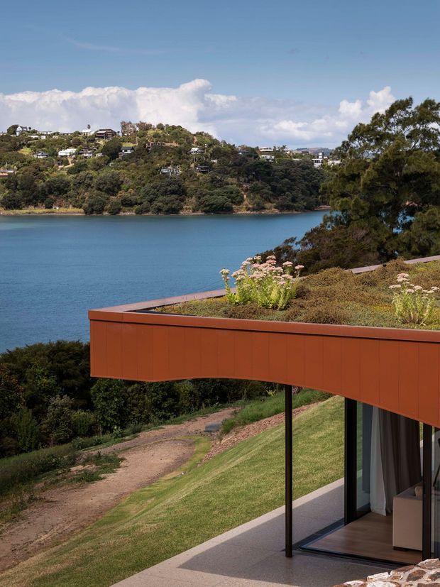 A contemporary waterfront house viewed from an elevated exterior terrace. The image features a projecting rust-orange clad roof or terrace edge with a planted green roof, slender dark supports, and extensive floor-to-ceiling glazing. Beyond the house is a wooded hillside with scattered homes across a calm blue inlet, while a sloping lawn and path descend in the foreground.