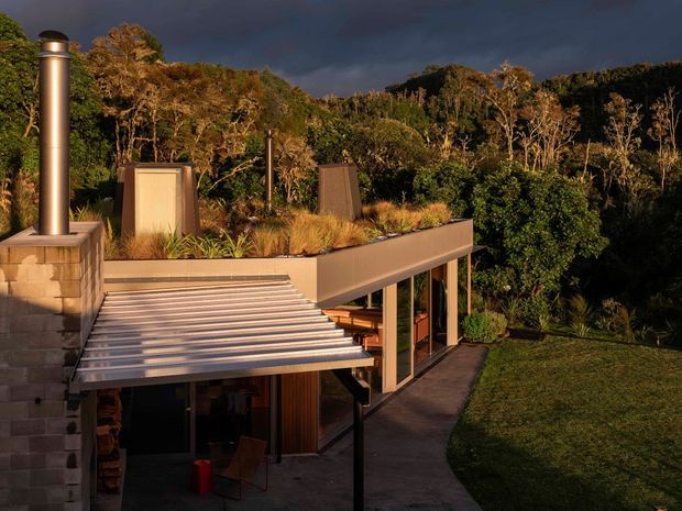 Exterior view of a contemporary, multi-level house integrated into a lush wooded landscape. The building features a flat green roof, broad projecting metal awnings, exposed structural posts, large glazed openings, and a tall metal flue or chimney. Warm low-angle sunlight contrasts with a dark, dramatic sky and dense native vegetation.