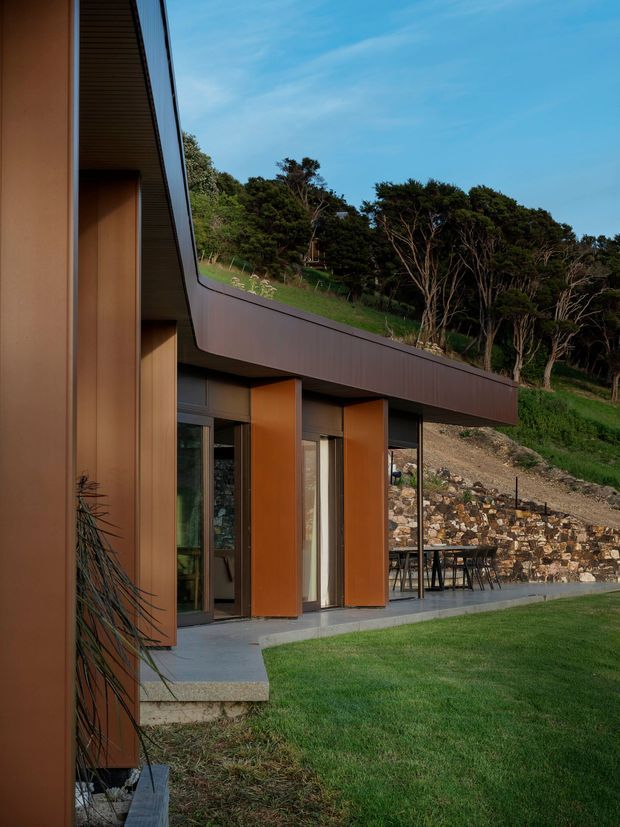 Exterior view of a contemporary hillside residence with a low, angular roofline and projecting brown metal-clad facade elements. Full-height glazing and glazed doors open onto a concrete terrace with outdoor dining furniture. A stone retaining wall, sloping landscaped site, and dense trees form the surrounding setting.