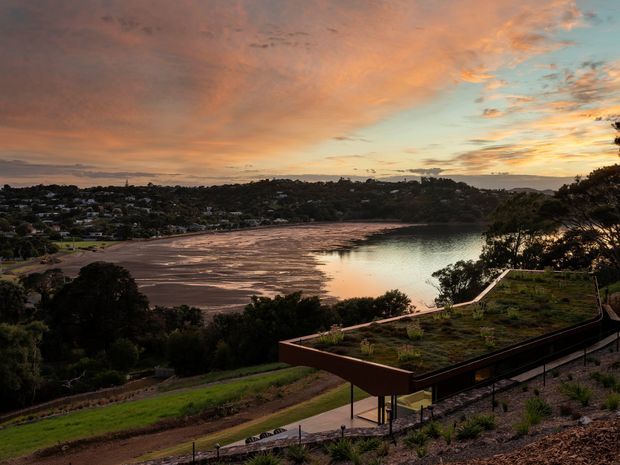 A dusk exterior landscape view overlooking a calm bay or inlet, with a suburban hillside settlement across the water. In the foreground, a contemporary landscaped property features a long angular terrace or viewing platform with dark metal railings, planted roof or garden areas, and a winding paved access path. The scene is illuminated by warm sunset light and dramatic orange-pink cloud cover.
