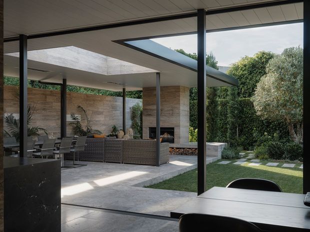 A sophisticated indoor-outdoor living and dining area opening onto a landscaped courtyard garden. The space features slim dark structural frames, expansive glazing, a pale panelled ceiling, large-format grey flooring, a long dining table, an outdoor dining setting, woven seating, a stone-clad wall with a built-in fireplace, and stepping stones through dense greenery.