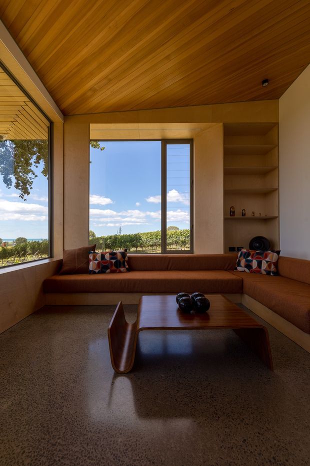 A warm, light-filled living room with expansive corner glazing overlooking a coastal landscape. The room features a built-in rust-brown sectional sofa with colourful geometric cushions, a low dark timber coffee table, polished concrete flooring, and extensive timber lining across the sloped ceiling and upper walls. Built-in open shelving is integrated beside the central window. The space has a strong mid-century modern character with natural materials and a compact, carefully composed layout.