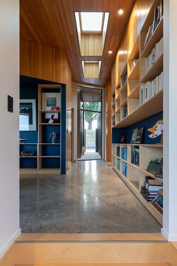 A narrow, warmly finished library hallway or passage with extensive built-in timber shelving filled with books and display items. The walls and sloped ceiling are clad in honey-toned timber panelling, with dark blue backing to portions of the shelving. Two large skylights bring natural light into the corridor, while polished speckled concrete flooring contrasts with the timber. A glazed door at the far end opens toward an outdoor area, and a step separates the foreground timber floor from the corridor.