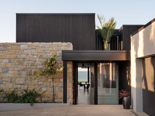 Contemporary multi-level residential facade with dark vertical cladding, pale rendered walls, and a substantial natural stone boundary wall. A recessed glazed entry opens through the house toward a visible body of water, while tropical planting and strong shadow patterns add a resort-like character. The image is taken from an exterior approach and is dominated by the building facade and entry composition.