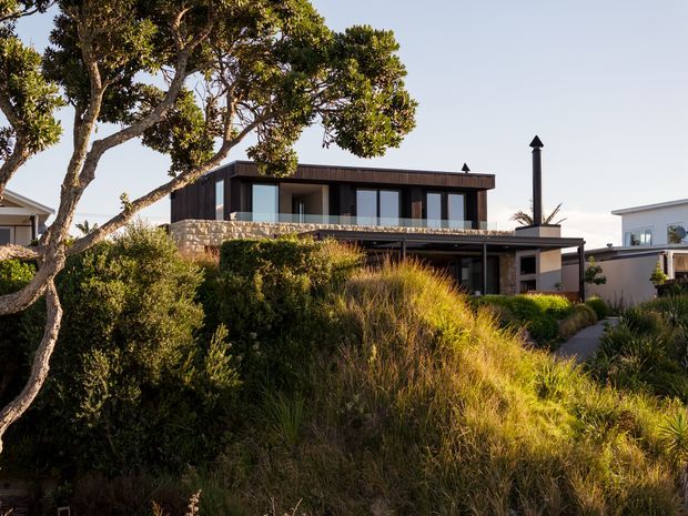 Exterior view of a contemporary two-storey coastal home set within established landscaped planting. The house features dark vertical cladding, broad horizontal rooflines, extensive glazing, a stone-clad balcony or retaining wall, and a sheltered lower-level entry terrace. Native grasses, shrubs, and mature trees dominate the foreground.