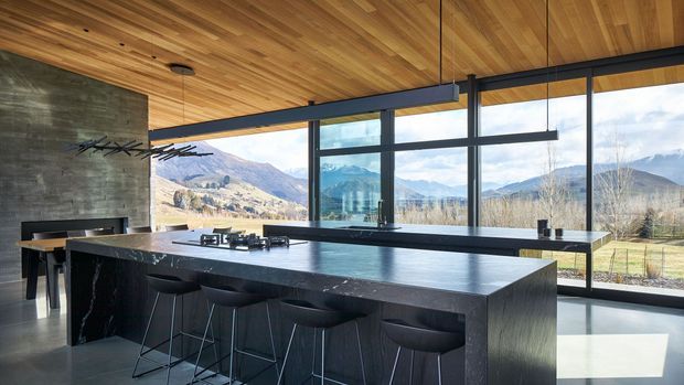 A contemporary kitchen with a large dark stone island and integrated seating, expansive black-framed glazing overlooking a mountainous landscape, and a warm timber-lined ceiling. A concrete feature wall, minimalist pendant lighting, and sleek dark cabinetry create a refined indoor-outdoor setting.