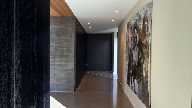 A long, contemporary hallway with dark timber wall panelling and cabinetry, a polished dark floor, a textured grey feature wall, recessed ceiling lights, and a large abstract artwork. Warm timber ceiling lining and contrasting pale walls add visual warmth to the otherwise dark, refined interior.