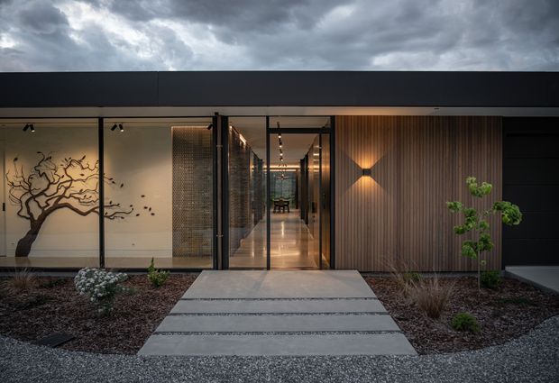 A dramatic contemporary single-storey building entrance viewed from an exterior landscaped approach. Broad concrete stepping slabs lead through gravel and drought-tolerant planting to a glazed entry framed in dark metal. Inside, a long illuminated corridor is visible, with a warm timber slat feature wall and wall lighting on the right, and an artistic branching wall mural on the left. A dark flat roofline and heavily overcast sky create a moody architectural presentation.