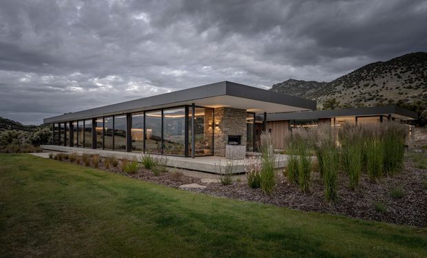A low-profile, single-storey contemporary residence set against rugged hills, photographed at dusk beneath dramatic cloud cover. The building features a broad flat roof, extensive floor-to-ceiling glazing with dark slender frames, stone-clad wall sections, warm interior lighting, and landscaped grounds with ornamental grasses and lawn.