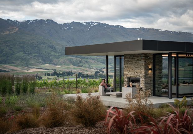 A contemporary single-storey pavilion-style home set among landscaped planting and a vineyard, with dramatic mountain ranges in the background. The building features a broad flat roof, extensive floor-to-ceiling glazing, slender dark frames, and a central stacked-stone fireplace wall. An outdoor terrace extends from the living area.
