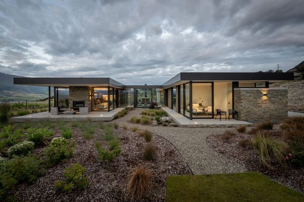 Wide exterior view of a low, contemporary single-storey residence arranged around a landscaped courtyard. Flat roof planes, extensive floor-to-ceiling glazing, dark slender frames, and warm illuminated interiors define the building. A natural stacked-stone wall anchors the right wing, while gravel paths, native-style planting, and grasses surround the outdoor spaces. Rolling hills are visible beyond the property beneath a dramatic overcast sky.