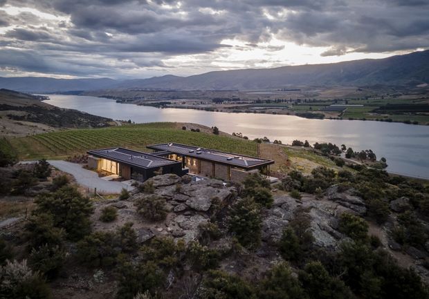 A low-profile modern residence set among rocky, scrub-covered terrain on a hillside overlooking a broad lake and surrounding mountainous valley. The building has dark flat roof planes, extensive glazing, and warm-toned exterior walls, with a winding driveway approaching it. Dramatic overcast evening light creates a muted, atmospheric landscape view.