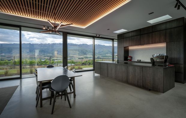 A contemporary open-plan kitchen and dining area with expansive floor-to-ceiling glazing overlooking a mountainous landscape. The space features dark wood kitchen cabinetry and island joinery, a smooth concrete floor, a large white dining table with dark timber chairs, and a striking slatted timber ceiling with integrated linear lighting. A skylight and minimalist track lighting add illumination, while the broad glazing creates a strong indoor-outdoor connection.