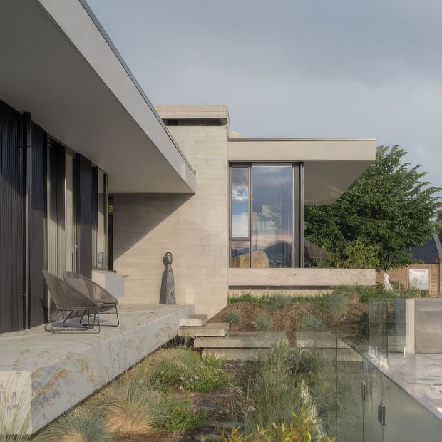 A refined contemporary residential exterior with strong horizontal cantilevered forms, pale stone or concrete walls, dark floor-to-ceiling glazing, and a sheltered terrace furnished with two lounge chairs. A landscaped garden and transparent glass pool or water-edge balustrade occupy the foreground, while the building’s geometric volumes and reflective windows dominate the composition.